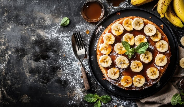 Bolo decorado com muitas fatias de banana sobre uma camada de calda de caramelo. Há uma folha de hortelã no centro para enfeitar. O bolo está em um prato preto, cercado por bananas maduras, talheres e açúcar de confeiteiro espalhado pela mesa.