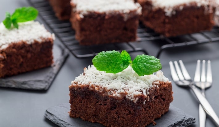 Fotografia de um quadrado de bolo de chocolate, coberto com coco ralado branco e decorado com uma folha de hortelã. O bolo está em uma tábua de ardósia escura, com mais pedaços ao fundo.