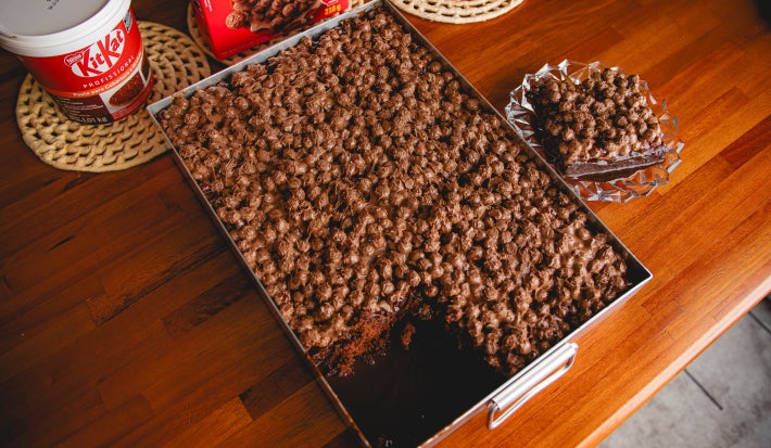 Fotografia mostra Bolo de chocolate úmido coberto com uma generosa camada de Pasta KITKAT® misturada ao NESCAU® Cereal, com brilho e textura crocante.