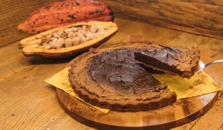 Fotografia em tons de marrom de uma bancada de madeira com uma tábua de madeira, sobre ela um paninho amarelo com uma torta de chocolate e uma espátula retirando uma fatia. Ao fundo um cacau.