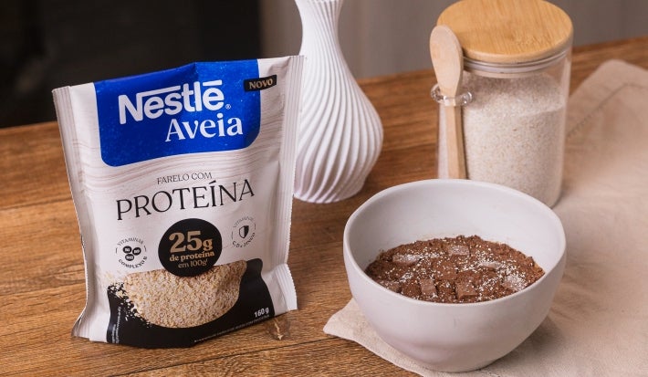 Receita de massa de brownie comestível servida em um pote branco sobre uma mesa de madeira decorada com um pote de aveia, um vaso e uma embalagem de nestlé aveia proteína