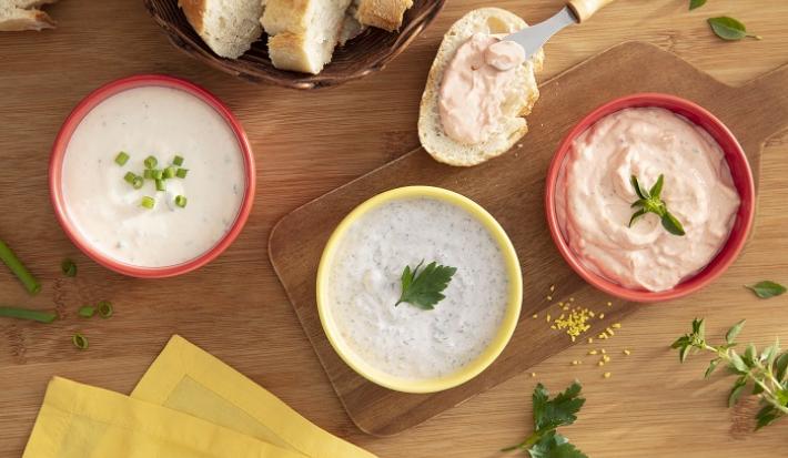 Foto da receita de trio de pates e ricota com maggi meu segredo servida em três porções com os diferentes patês em potinhos vermelhos e amarelos. Tudo isso sobre uma mesa de madeira decorada com pães e ervas