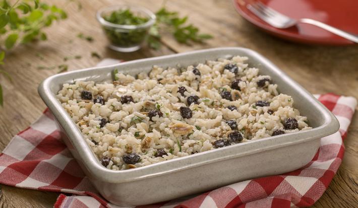 fotografia em tons de bege, branco e vermelho de uma bancada bege vista de frente. Contém um pano xadrez nas cores branco e vermelho com um recipiente quadrado branco com o arroz e passas. Ao fundo um prato vermelho com uma colher.