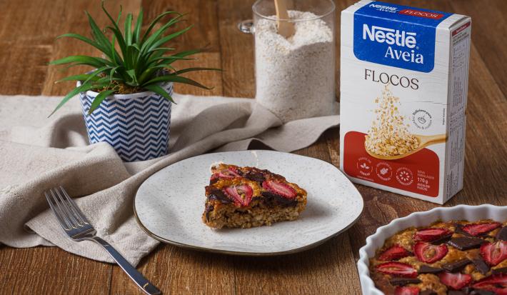 Fotografia de um prato branco com borda preta, ao lado de uma embalagem de aveia flocos, sobre uma mesa de madeira. Sobre o prato tem uma fatia do bolo de aveia com morango e chocolate.