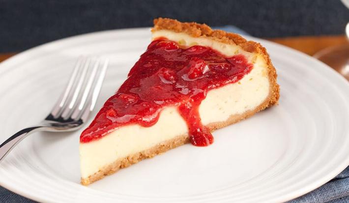 Fotografia em tons de branco e vermelho, no centro na foto está uma fatia da torta coberta com geleia de morango, ao lado está um garfo e embaixo do prato um guardanapo azul.