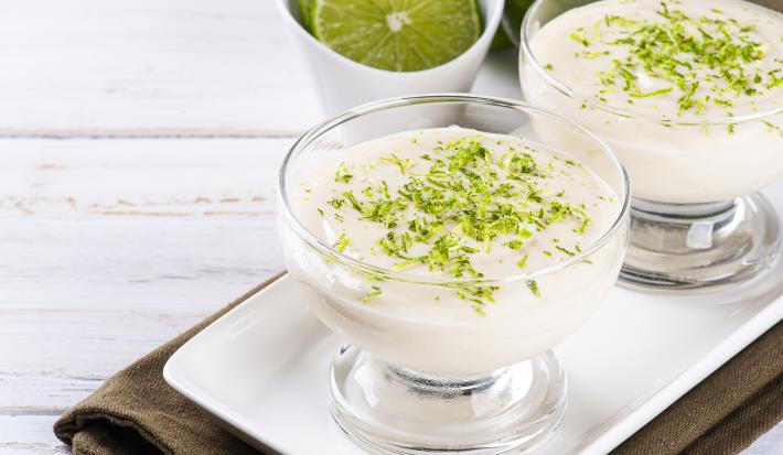 Foto aproximada da receita de Mousse de Limão em tom claro, em um pote redondo de vidro e decorado com raspas de limão. Tudo sobre um prato branco e um tecido marrom escuro. No rodapé há uma faixa verde com dizeres.