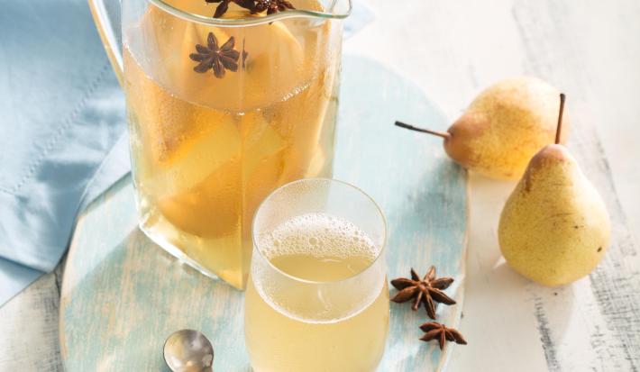 Fotografia em tons de azul em uma bancada de madeira cinza clara, um pano azul, uma bandeja de madeira azul mesclada com uma jarra de vidro e o refresco de pera e erva-doce dentro dele. Ao lado, um copo de vidro com a bebida, peras e anis-estrelado.