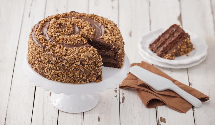 Fotografia em tons de marrom em uma mesa de madeira com um suporte para bolo branco ao centro e o bolo crocante de chocolate em cima. Ao lado, um guardanapo de pano marrom com uma espátula de bolo branco apoiada e um pratinho branco com uma fatia do bolo.