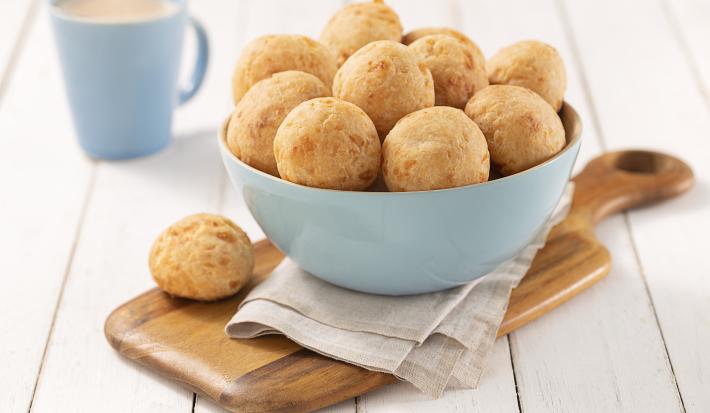 Foto em tons de bege com os pães de queijo em um bowl azul claro sobre um paninho cinza em cima de uma tábua de madeira, com uma unidade à frente do bowl para decorar. No fundo como decoração há uma xícara azul de café.