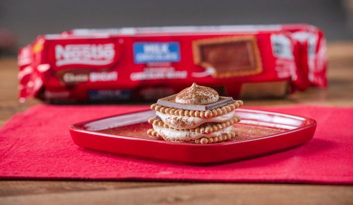 Fotografia de um tiramissu em camadas de creme e choco biscuit sobre um prato quadrado vermelho e raso com cacau em pó polvilhado. O prato está apoiado em um pano vermelho, e ao fundo, tem um pacote de choco biscuit.