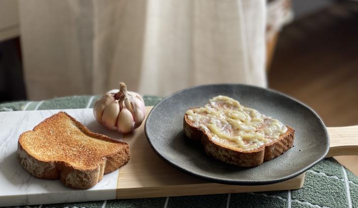 Foto de um prato escuro redondo com uma fatia de pão com a manteiga de alho por cima. Ao lado esquerdo dele há outra fatia de pão torrada e um dente de alho inteiro ao fundo.