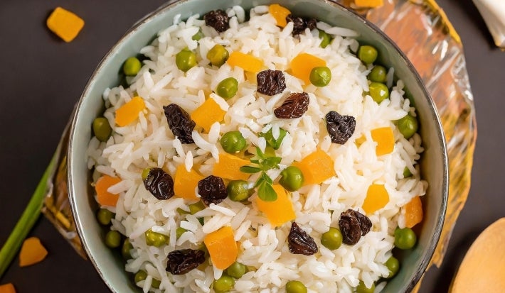 Imagem de cima de uma tigela com arroz colorido com passas, ervilhas e cenouras picadas.