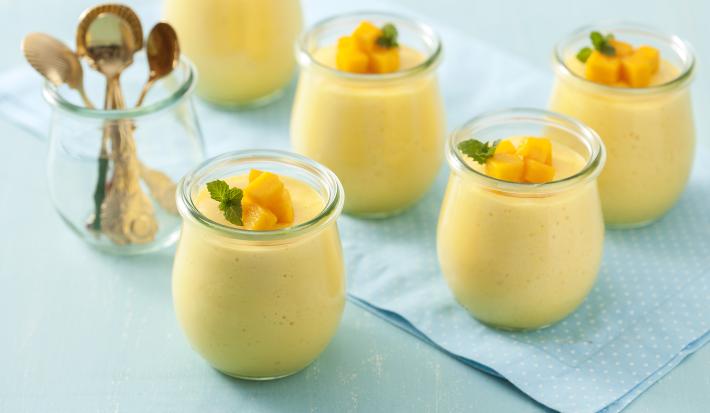 Fotografia em tons de azul e amarelo de uma bancada azul com um paninho azul com bolinhas brancas, um pote transparente com quatro colheres douradas e cinco potes transparentes com a mousse de manga, decorados com cubinhos de manga e uma folha de hortelã.