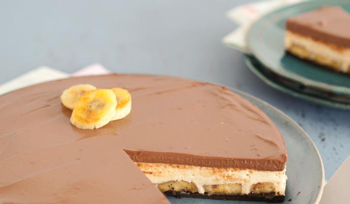 Fotografia em tons azul em uma mesa de madeira azul, com um pano branco com rosa florido, um prato azul raso redondo com a torta mousse de doce de leite e banana.
