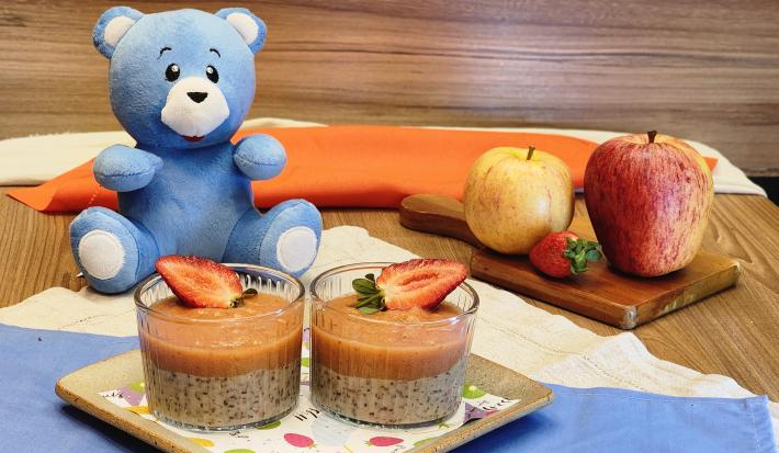 Fotografia de dois copos pequenos de vidro com pudim de pera com morango, e por cima de cada um, tem uma metade de morango para decorar. Os recipientes estão sobre um apoio quadrado. Ao fundo, tem o urso azul de mucilon e as frutas usadas na receita.