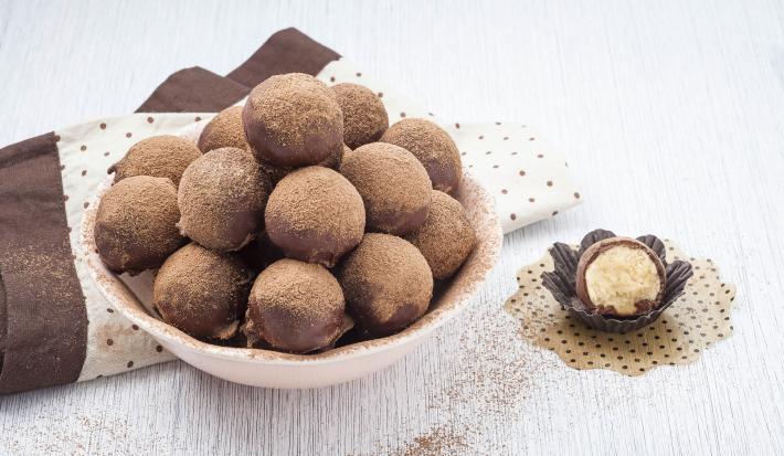 foto em tons de marrom, branco e rosa de uma bancada vista de cima. Contém um recipiente redondo cor rosa com trufas de chocolate e abaixo um pano marrom e um pano branco com bolinhas marrom. Ao lado uma trufa pela metade em uma forminha preta