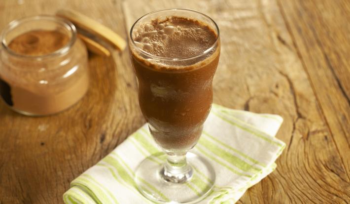 Fotografia em tons de marrom e branco de uma bancada de madeira com um paninho branco e verde, sobre ele um copo de vidro com o smoothie. Ao fundo um recipiente de vidro com cacau em pó.