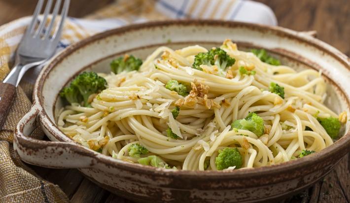 Fotografia em tons de verde em uma bancada de madeira com um pano xadrez em bege e azul, um recipiente marrom com o macarrão alho e óleo com brócolis.