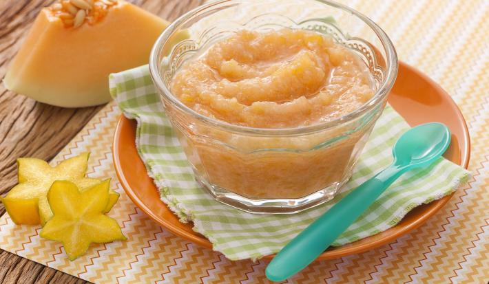 Fotografia em tons de laranja em uma bancada de madeira com um paninho xadrez verde e um potinho de vidro em cima com o mingauzinho de melão e carambola. Ao lado, um pedaço de um de melão e duas fatias cortadas de carambola.