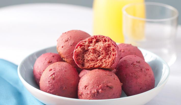 Fotografia em tons de azul e rosa em uma mesa branca com um pano azul ao lado e um prato branco fundo ao centro com os pães de beterraba. Ao fundo, uma jarra com suco de laranja e um copo de vidro.