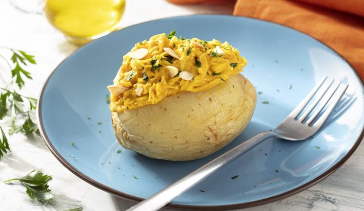 Fotografia em tons de amarelo em uma bancada de madeira de cor branca. Ao centro, um prato azul contendo a batata com um garfo ao lado. Ao fundo, há um pano laranja, um pote de azeite e algumas ervas espalhadas.