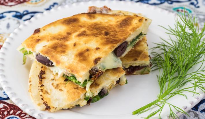 Fotografia em tons de branco com um prato branco ao centro. Em cima do prato existe uma receita de quesadilla de queijo com espinafre.