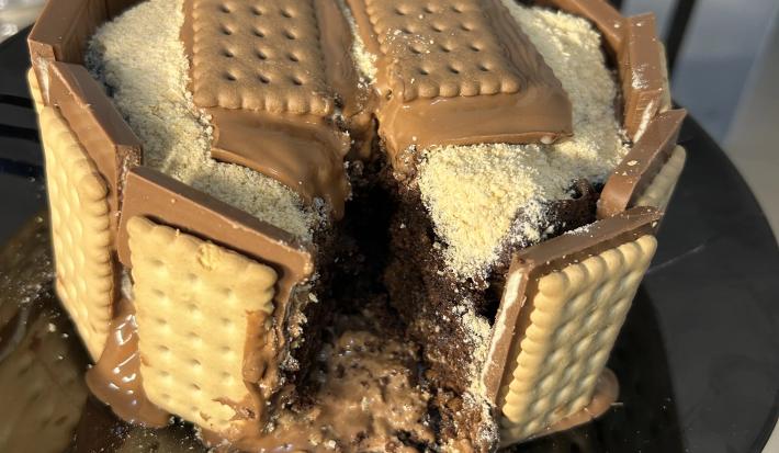 Fotografia de um bolo de chocolate com brigadeiro e farinha láctea polvilhada. Ao redor do bolo tem chocotrio de amendoim grudado. O bolo está sobre um prato de vidro preto raso.
