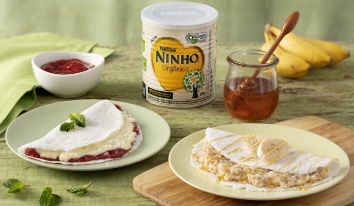 Fotografia em tons de verde de uma bancada verde com um prato redondo branco, sobre ele uma tapioca. Ao lado uma tábua de madeira com um prato redondo e uma tapioca. Ao fundo uma lata de ninho, um cacho de bananas e dois recipiente com mel e goiabada