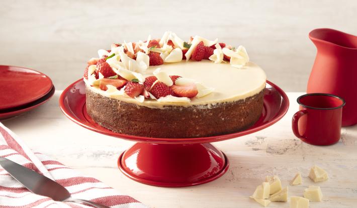 Fotografia em tons de vermelho em uma bancada bege com um paninho listrado branco e vermelho e ao centro um suporte vermelho com o bolo de três leites com ganache de chocolate branco. Ao lado, pedaços de chocolate branco, uma caneca e uma jarra vermelhas.
