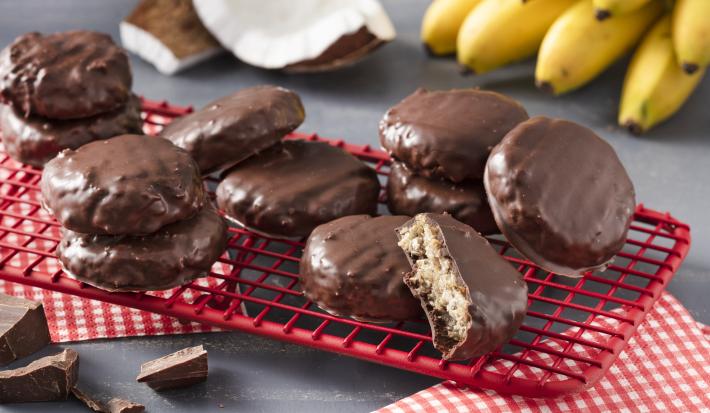 Fotografia em tons de vermelho em uma bancada cinza com um apoiador vermelho com os biscoitos de aveia, cacau e coco. Ao fundo, um cacho de bananas e pedaços de coco.