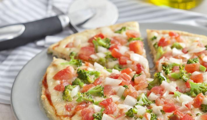 fotografia em tons de branco de uma bancada vista de frente, contém um prato redondo e branco com uma pizza com pedaços de tomates e brócolis ao fundo uma jarra com azeite.