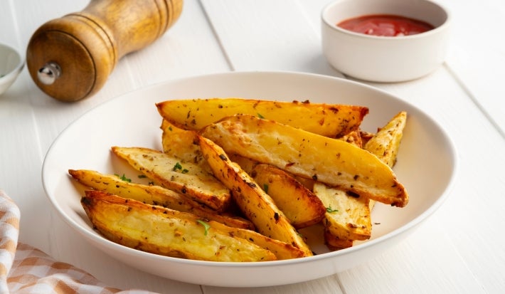 Fotografia em tons claros de um recipiente branco médio, fundo, sobre uma mesa de madeira branca. Dentro do recipiente tem batatas assadas e temperadas, cortadas em meia lua. Ao lado do recipiente tem temperos e ketchup em potinhos brancos.
