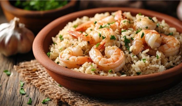 Em cima de uma mesa, prato de porcelana redondo com arroz branco soltinho e camarões, decorado com cheiro-verde.