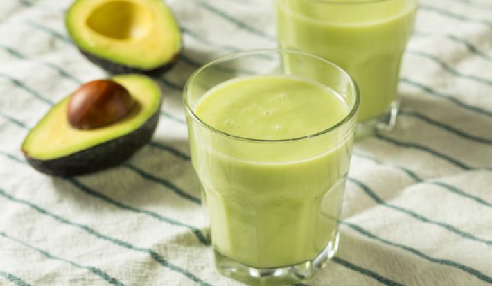 Fotografia de dois copos de vidro com vitamina de abacate. Ao fundo, ao lado do segundo copo, tem um abacate cortado ao meio, sobre uma toalha de mesa branca com listras verdes.