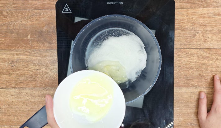Uma mão despeja claras de ovos dentro de uma panela com açúcar no fundo sobre um fogão portátil preto.
