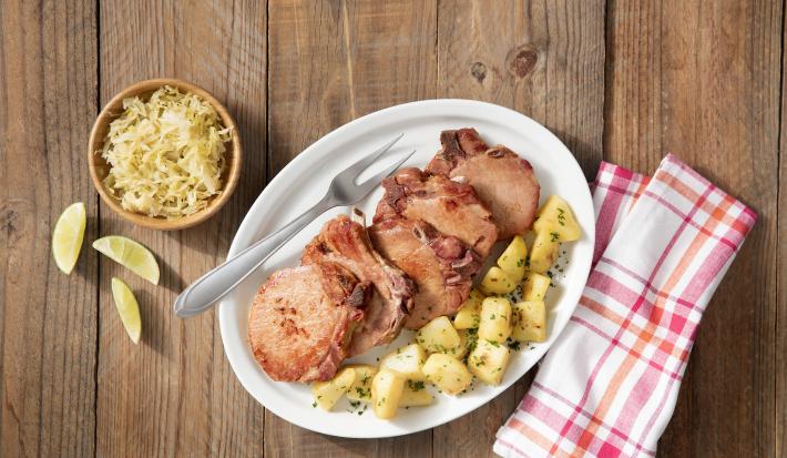 Fotografia em tons de branco de marrom, com bancada de madeira vista de cima, ao centro travessa branca contendo bisteca suína, batata picada e um garfo, no entorno potinho com repolho e fatias de limão e guardanapo quadriculado em branco e vermelho.