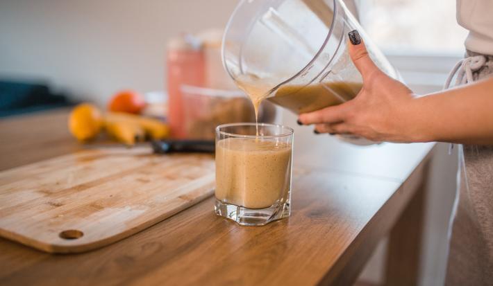 Fotografia de uma moça colocando uma bebida de banana e maçã feita no liquidificador em um copo de vidro pequeno, que está sobre uma mesa de madeira. Ao fundo, uma tábua de madeira e frutas, como bananas mamão e maçã.