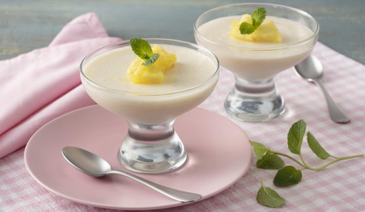 Fotografia em tons de rosa e amarelo de uma bancada, ao centro um paninho quadriculado rosa e branco, um prato rosa com uma taça com o mousse e pedacinhos de abacaxi e folha de hortelã. Ao lado uma taça igual e folhas de hortelã.
