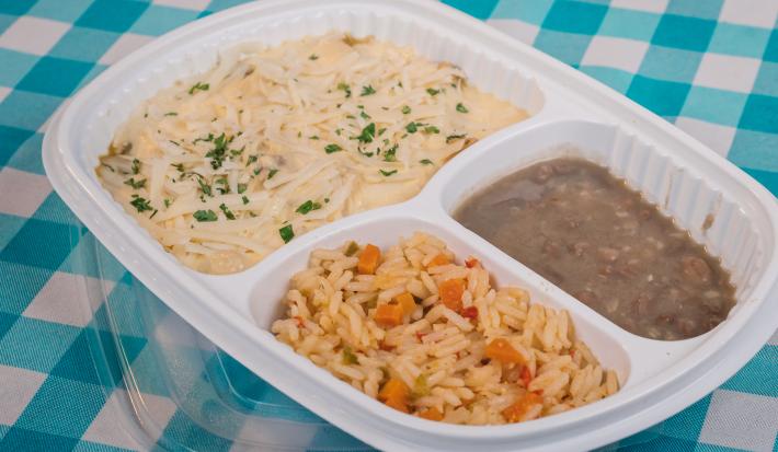 Foto aproximada de uma marmita branca de plástico com três divisões, uma com o frango com creme de milho, outra com arroz e outra com feijão. Ela está sobre um pano azul e branco quadriculado