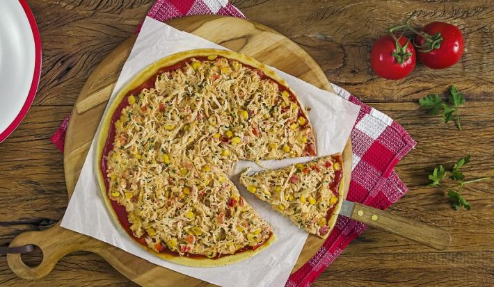 Fotografia em tons de marrom de uma bancada de madeira com uma tábua de madeira, sobre ela um paninho branco e um xadrez com uma pizza de frango e uma espátula retirando uma fatia. Ai lado dois tomatinhos e um prato branco com bordas vermelhas.