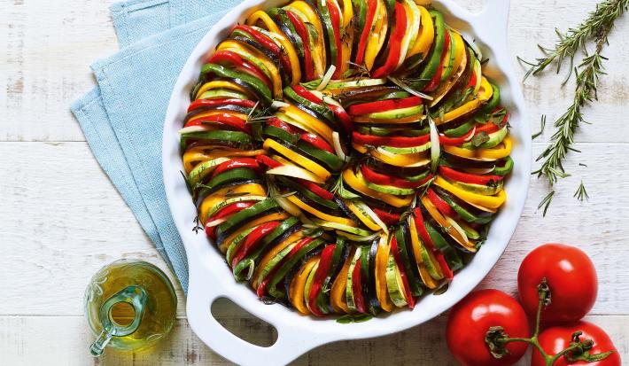 Fotografia em tons de verde, amarelo e vermelho de uma bancada branca com três tomates, uma jarrinha de azeite, ramos de alecrim e um recipiente branco redondo com alças com camadas intercaladas de abobrinha, berinjela e pimentões.