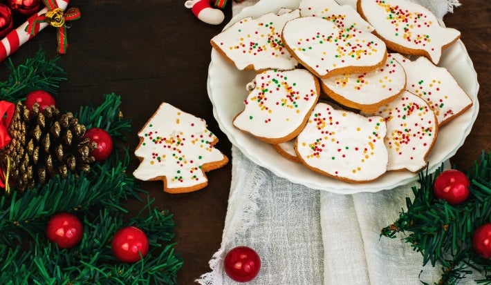 Em cima de uma mesa, prato redondo com biscoitos em forma de árvore e diversas formas, decorados com glacê branco e confeitos coloridos de bolinha.