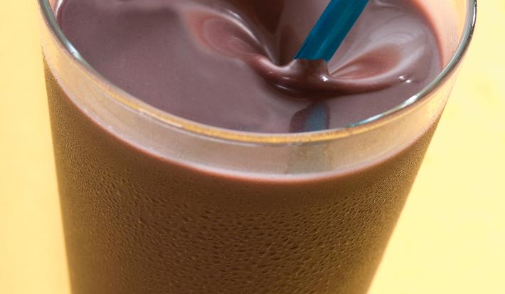 Fotografia em tons de amarelo em uma mesa de madeira amarela, com um copo de vidro alto com a vitamina de açaí dentro dele.