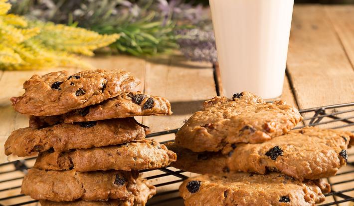 Fotografia em tons de marrom de uma bancada marrom com uma grade preta com cookies. Ao fundo ramos de flores e um copo de vidro com leite.
