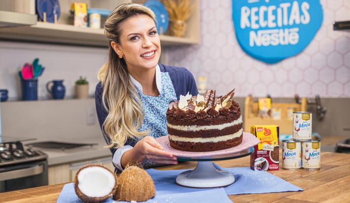 Imagem da confeiteira e influenciadora Beca Milano segurando a receita de bolo Naked de Prestígio atrás de uma bancada de cozinha, com pedaços de coco fresco e alguns produtos do lado direito