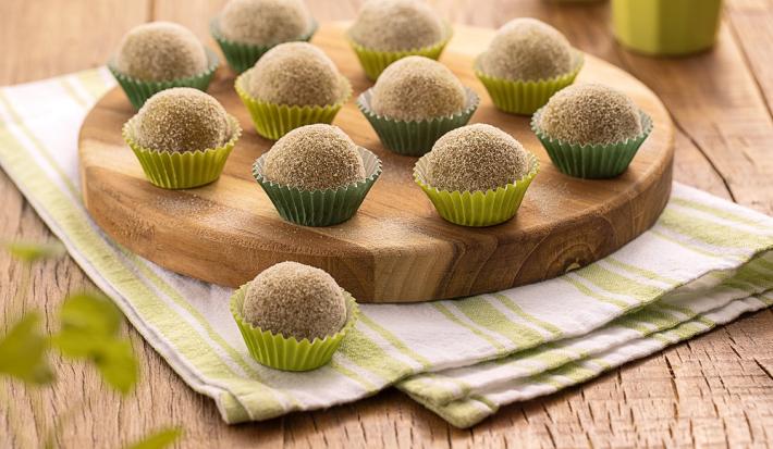 Foto da receita de Brigadeiro de Batata Doce. Na foto, observa-se brigadeiros em forminas nos tons de verde, sobre uma tábua redonda de madeira e um guardanapo listrado.