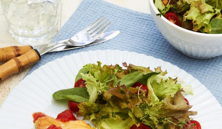Fotografia em tons de azul em uma bancada de madeira clara, um pano azul, um prato branco redondo raso com o frango à parmegiana e uma salada verde em cima dele. Ao lado, talher, um copo com água e rodelas de limão e um potinho com mais salada.