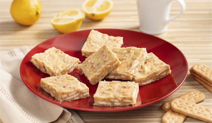 Fotografia em tons de amarelo, vermelho e branco de um paninho listrado amarelo e branco, sobre ele um prato redondo vermelho com palha italiana. Ao lado um paninho branco e biscoitos tostines maizena. Ao fundo uma xícara branca e dois limões.
