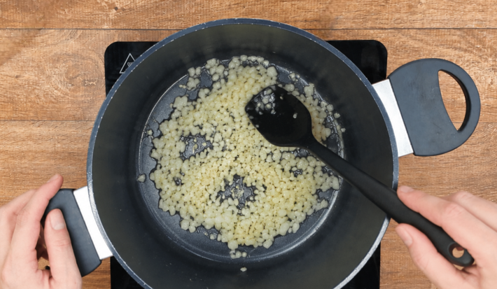 Cebola e alho refogando em panela sobre fogão portátil preto.