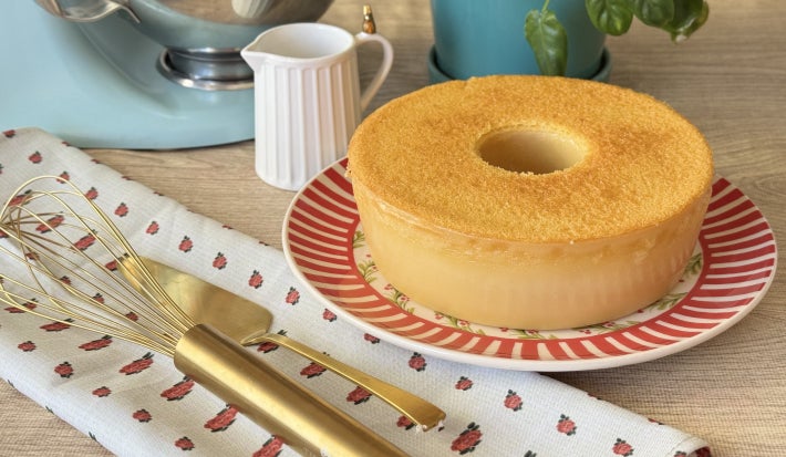 Foto da receita de bolo mole cearense, servido em um prato colorido, sobre uma mesa de café da tarde, com talheres dourados, uma batedeira azul ao fundo